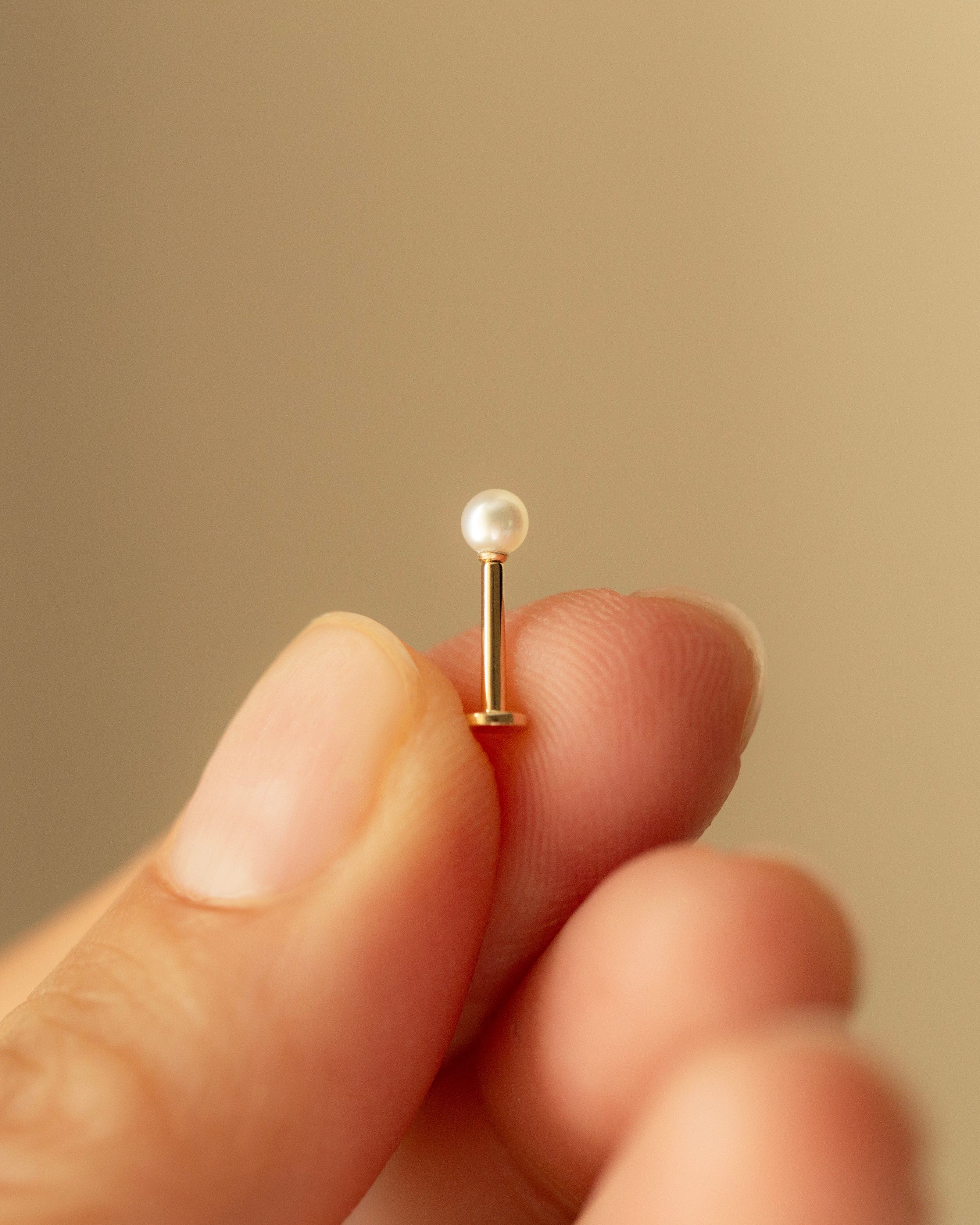 Close-up of a small pearl earring held between fingers against a beige background
