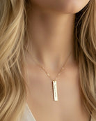 Gold necklace with a vertical bar pendant on a person with blonde hair.
