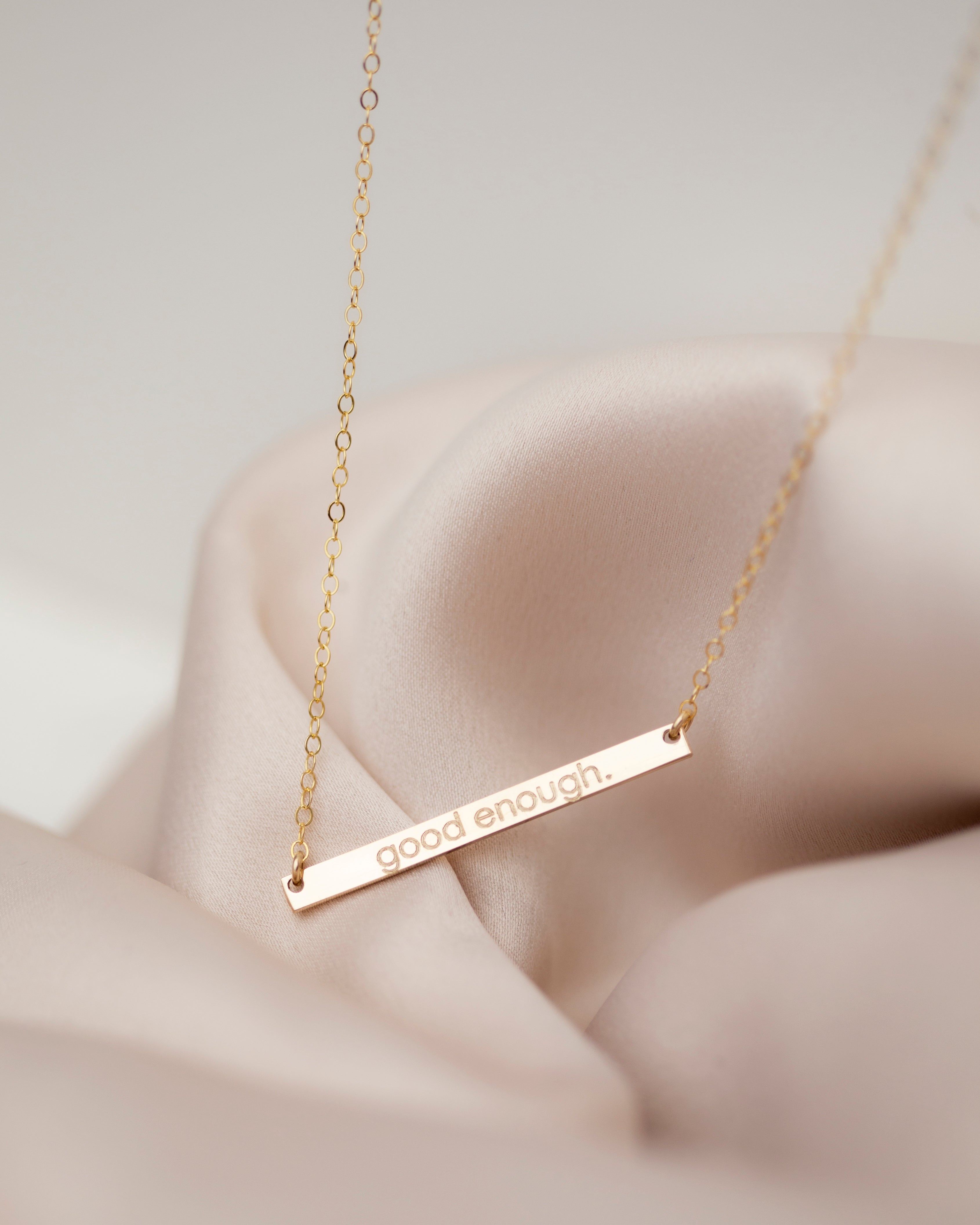 A gold-filled skinny name bar plate necklace laid out on a silky surface, with the bar featuring engraved personalized text.